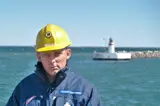 Man in blue jacket and yellow helmet in front of lighthouse