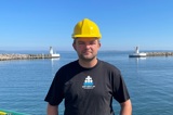 Man in black shirt and yellow helmet in front of lighthouse