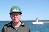 Man with glasses and gray shirt and green helmet in front of lighthouse